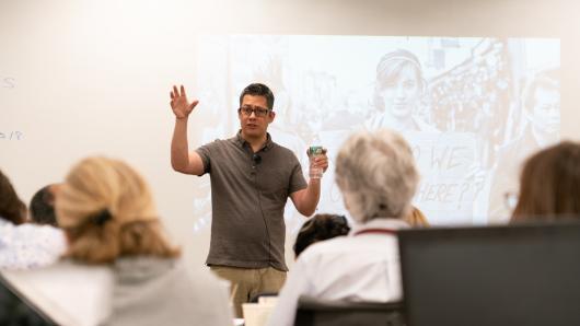 A man speaks to a group in front of a projected image of a woman holding a sign.