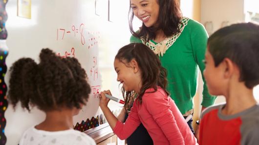 Smiling teacher stands with three young children at a dry-erase board working to solve math problems