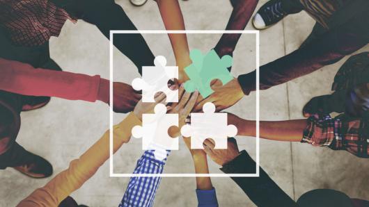 a racially/ethnically diverse group of people place their hands together beneath a graphic of puzzle pieces coming together