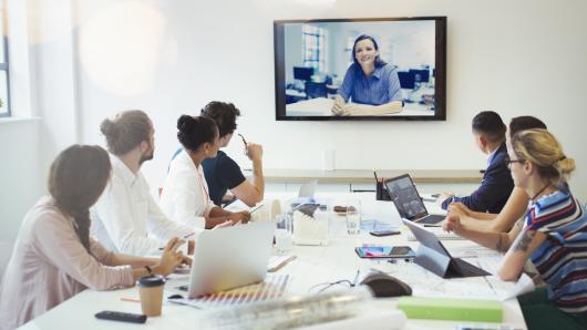 Group of 7 adults at conference table with computer facing virtual person on large screen